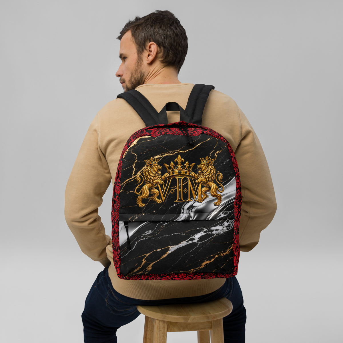 A man wearing a beige sweatshirt and dark jeans sits on a wooden stool, viewed from behind, wearing a black backpack with a red patterned border, gold lion-and-crown monogram, and a black-and-white marble print.