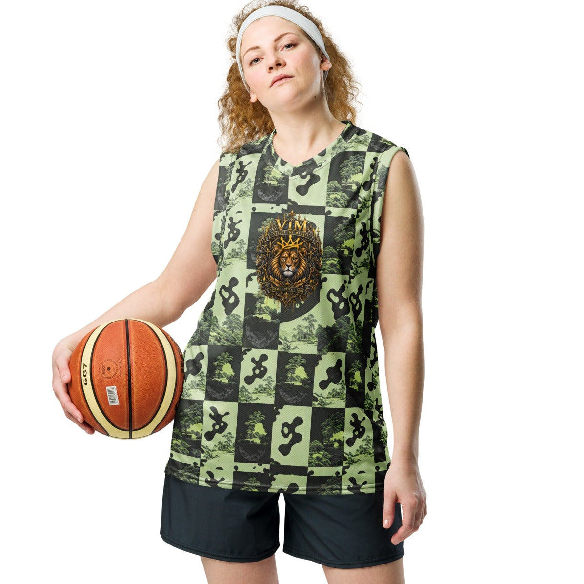 A woman modeling a sleeveless green and black patchwork basketball jersey with a central graphic, paired with black athletic shorts, white socks, and white sneakers, while holding a basketball against a plain white background.