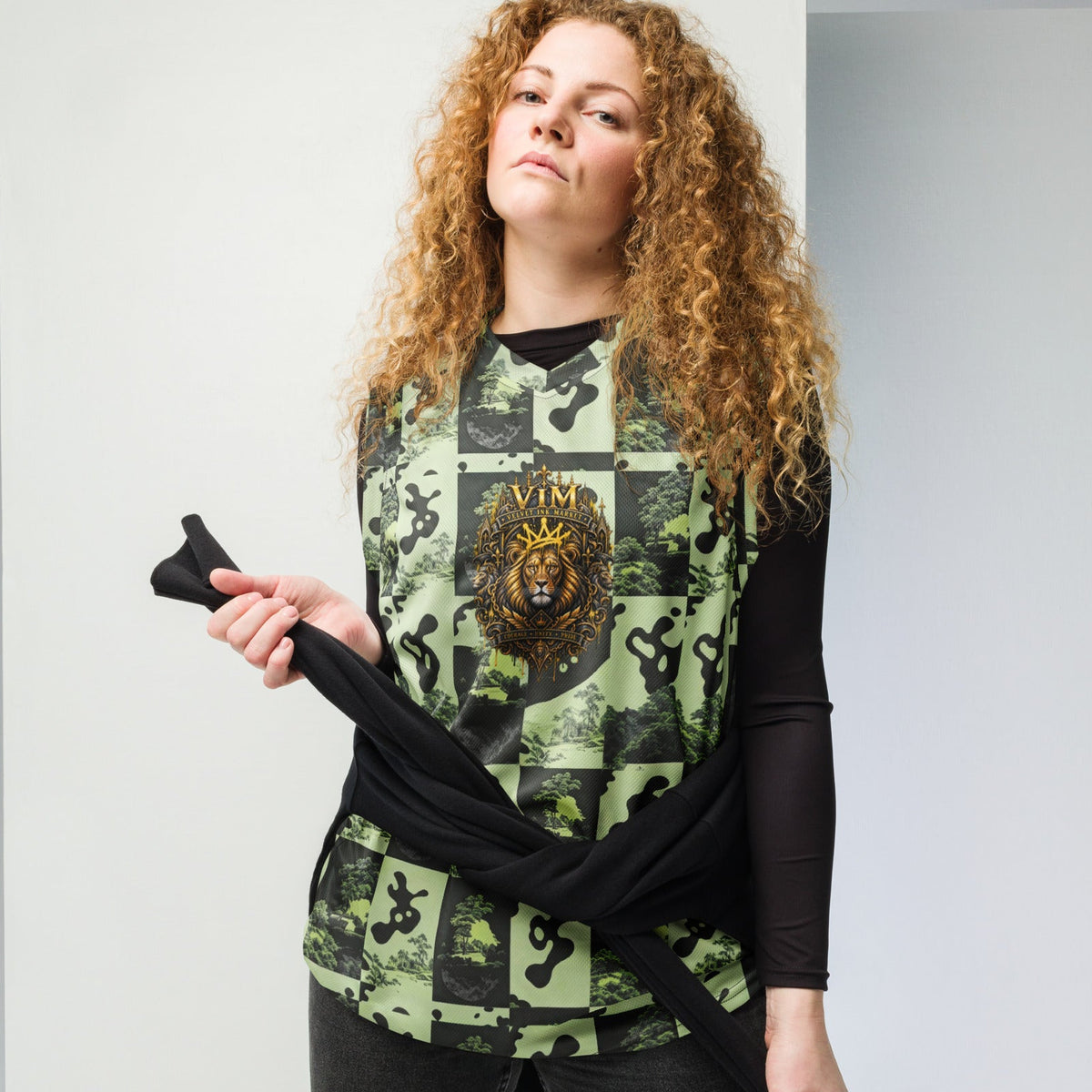 A woman with curly light brown hair models a sleeveless black top featuring a green and black collage print with a central gold lion emblem and the word 'VIVI'. She is holding a matching black cardigan draped over her arm, set against a minimalist light gray studio background.