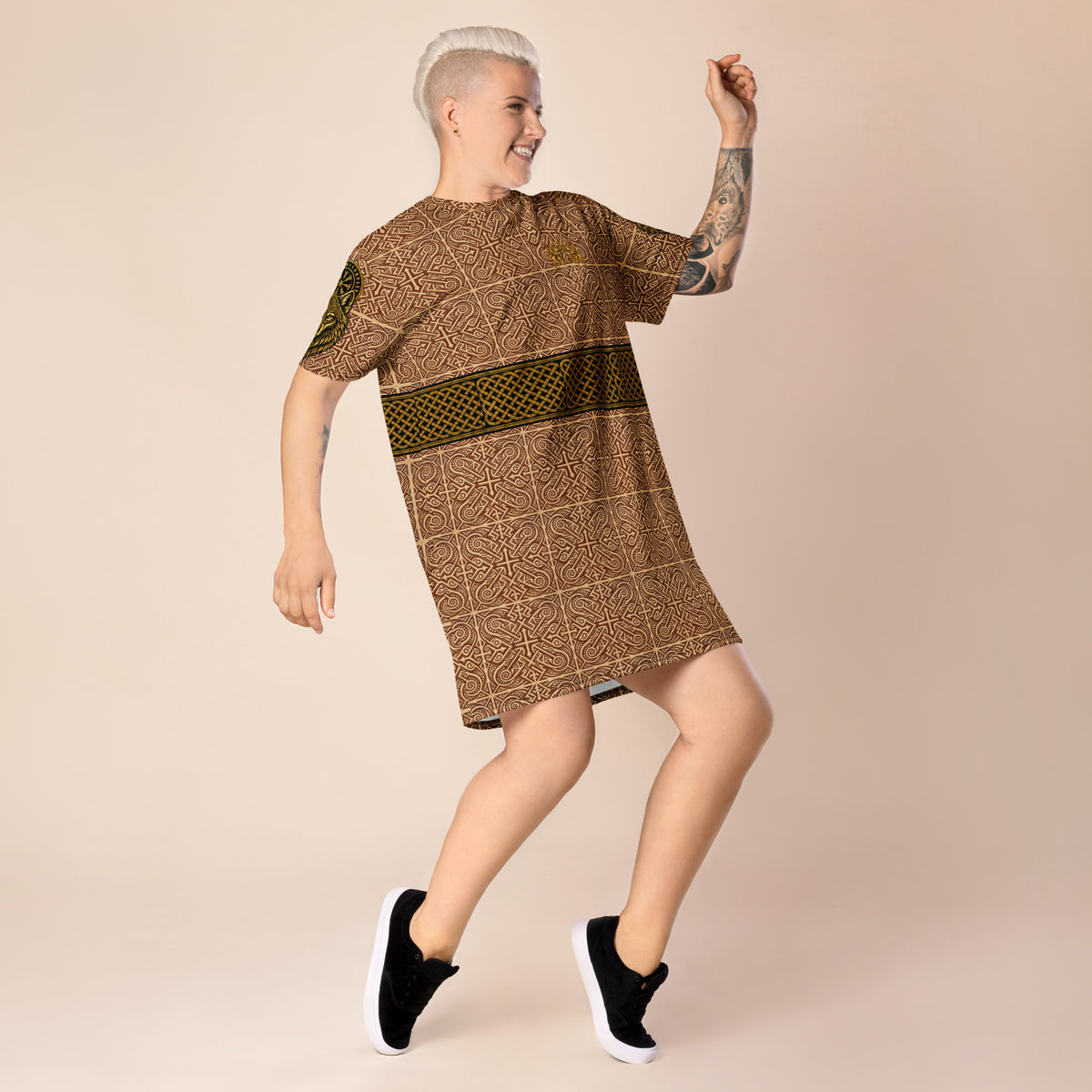 A person with short platinum blonde hair and tattoos is wearing a brown and olive green patterned short-sleeve t-shirt dress, posing dynamically against a solid beige studio background.