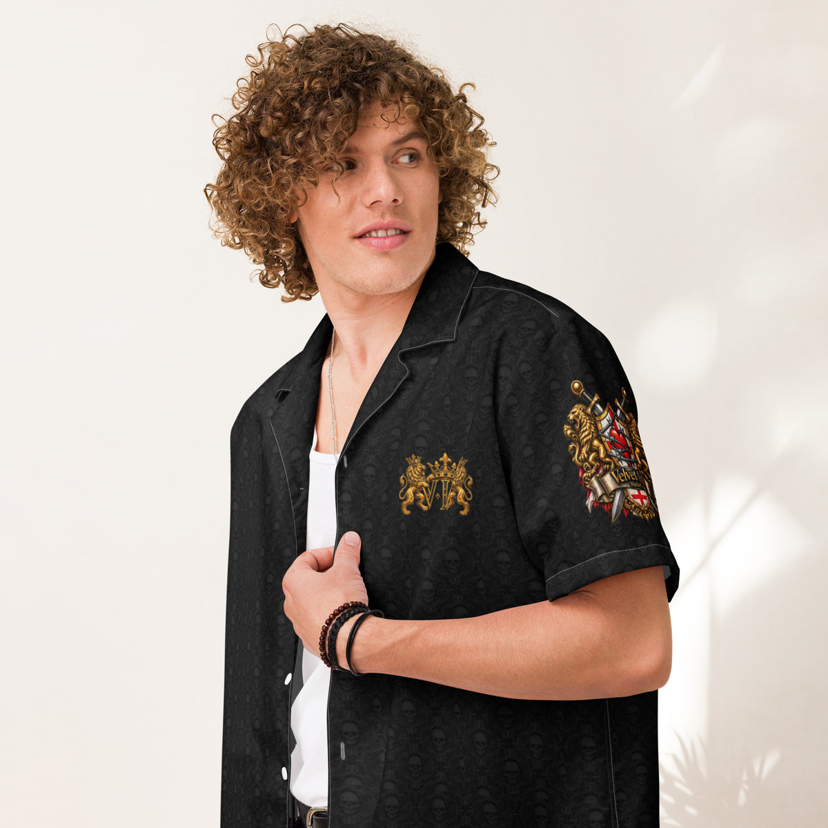 A man with curly brown hair wearing a black short-sleeve button-up shirt with embroidered gold and red crest designs on the chest and sleeve, styled over a white undershirt against a warm off-white background.