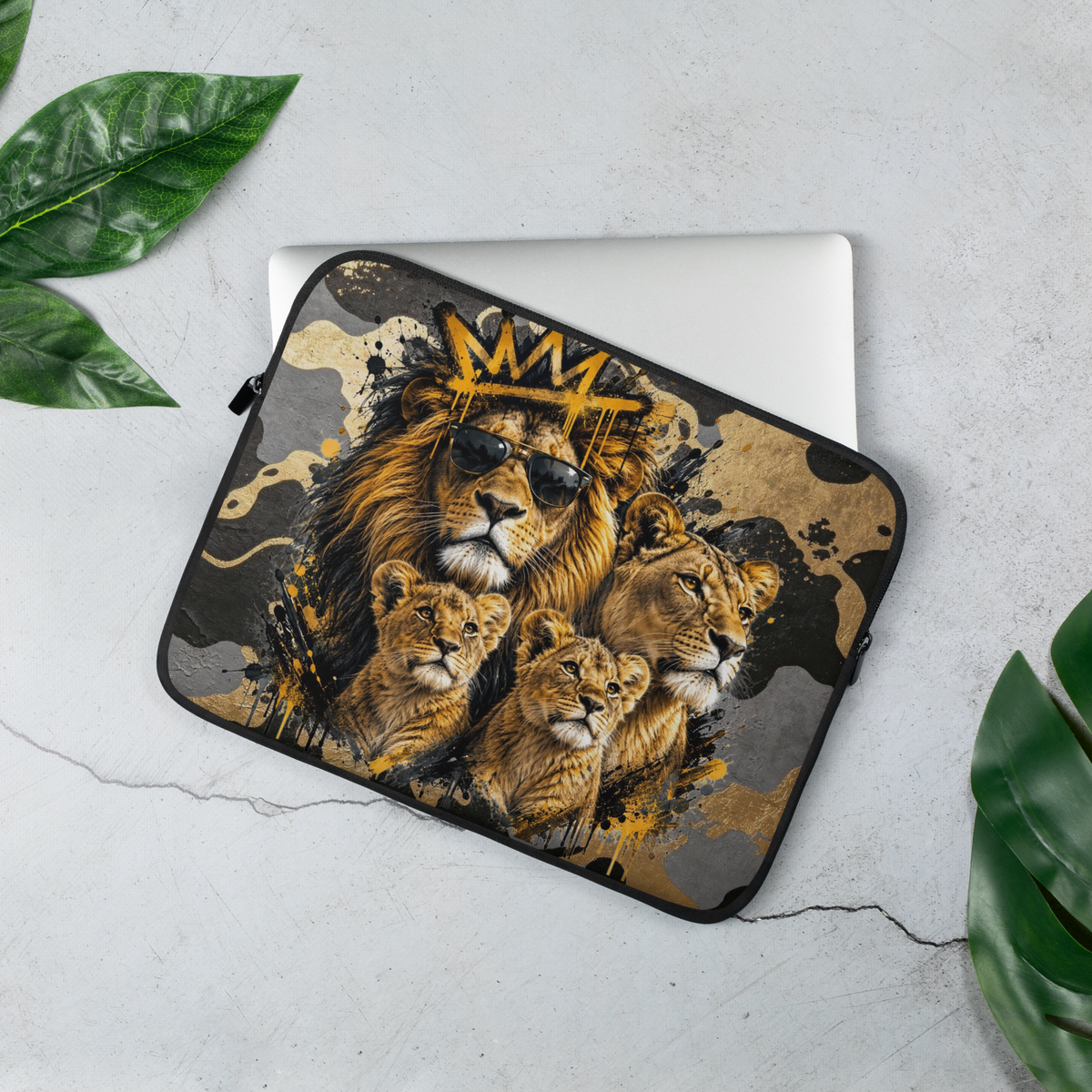 A laptop sleeve featuring a graphic print of lions and lion cubs with a gold crown, shown on a silver laptop and styled on a light gray concrete surface with green leaves.